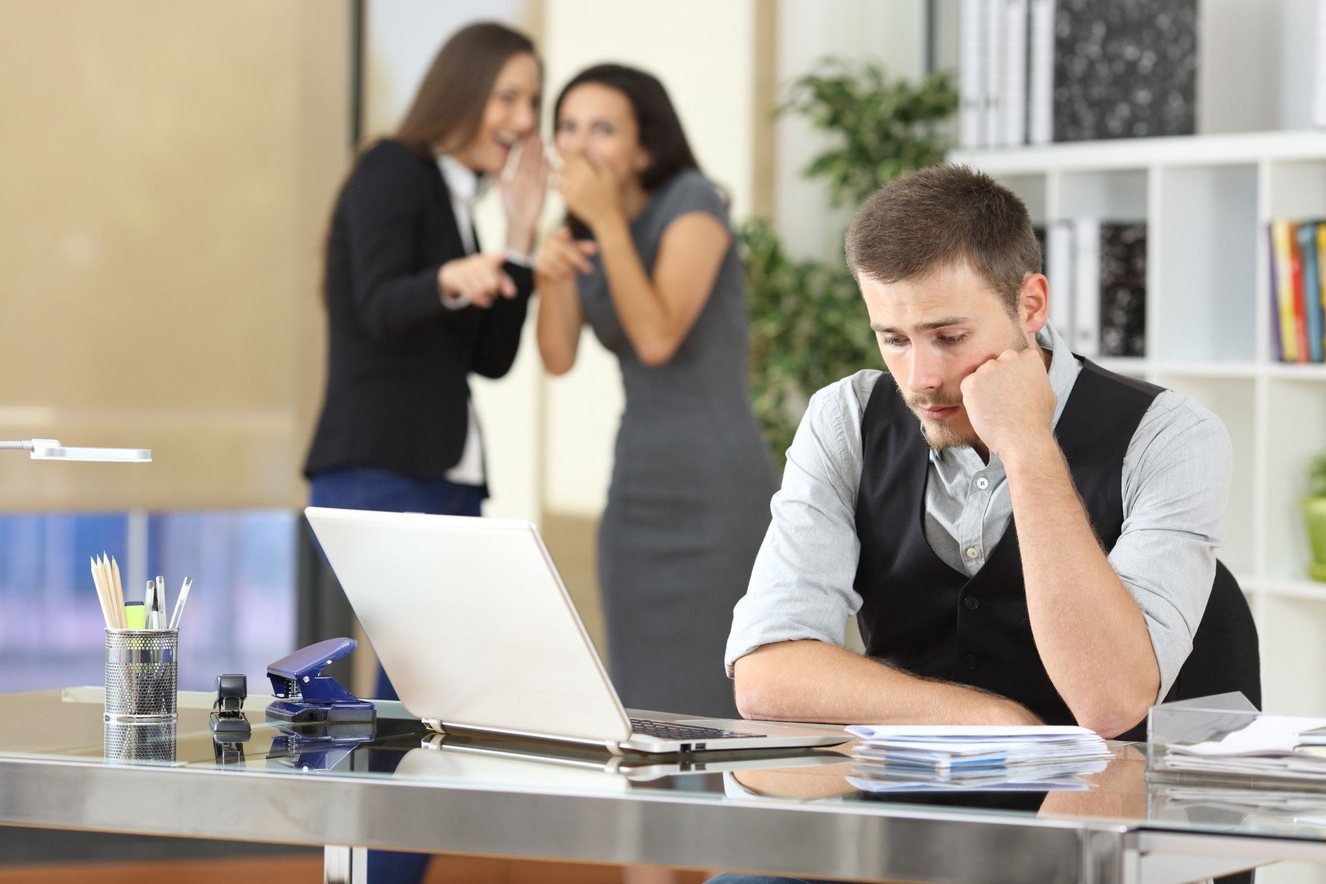 A man is sitting at a desk with a laptop and two women are whispering in the background.